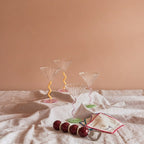 Inhoud van geschenkbox Social Gathering: vier &klevering Curve coupe cocktailglazen in caramel en roze, geborduurde Zigzag cocktailservetten en de Addison Ross Bobbin flesopener in Cherry & Cappuccino